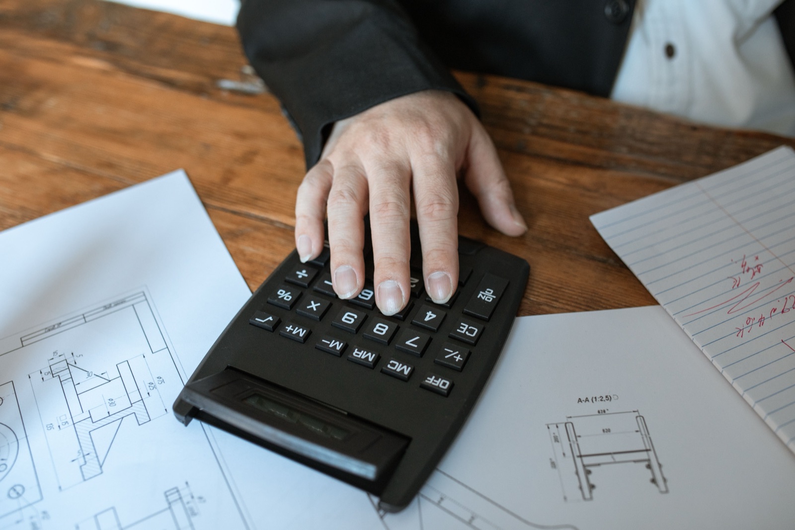 A hand on a desk calculator beside technical drawings and handwritten notes